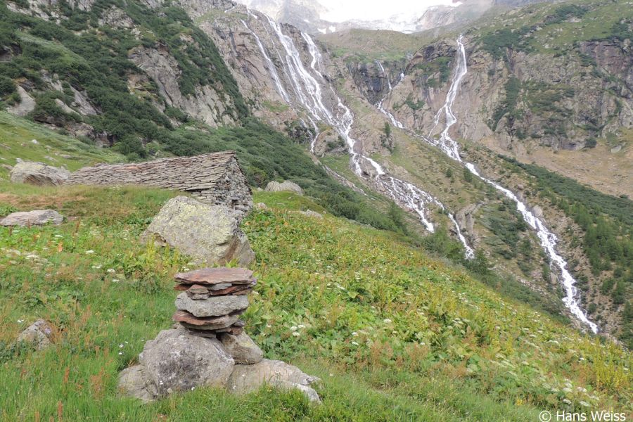 Alpine Wässerfälle stürzen ein steiles Gefälle hinunter. Im Vordergrund sind gestapelte Steine.